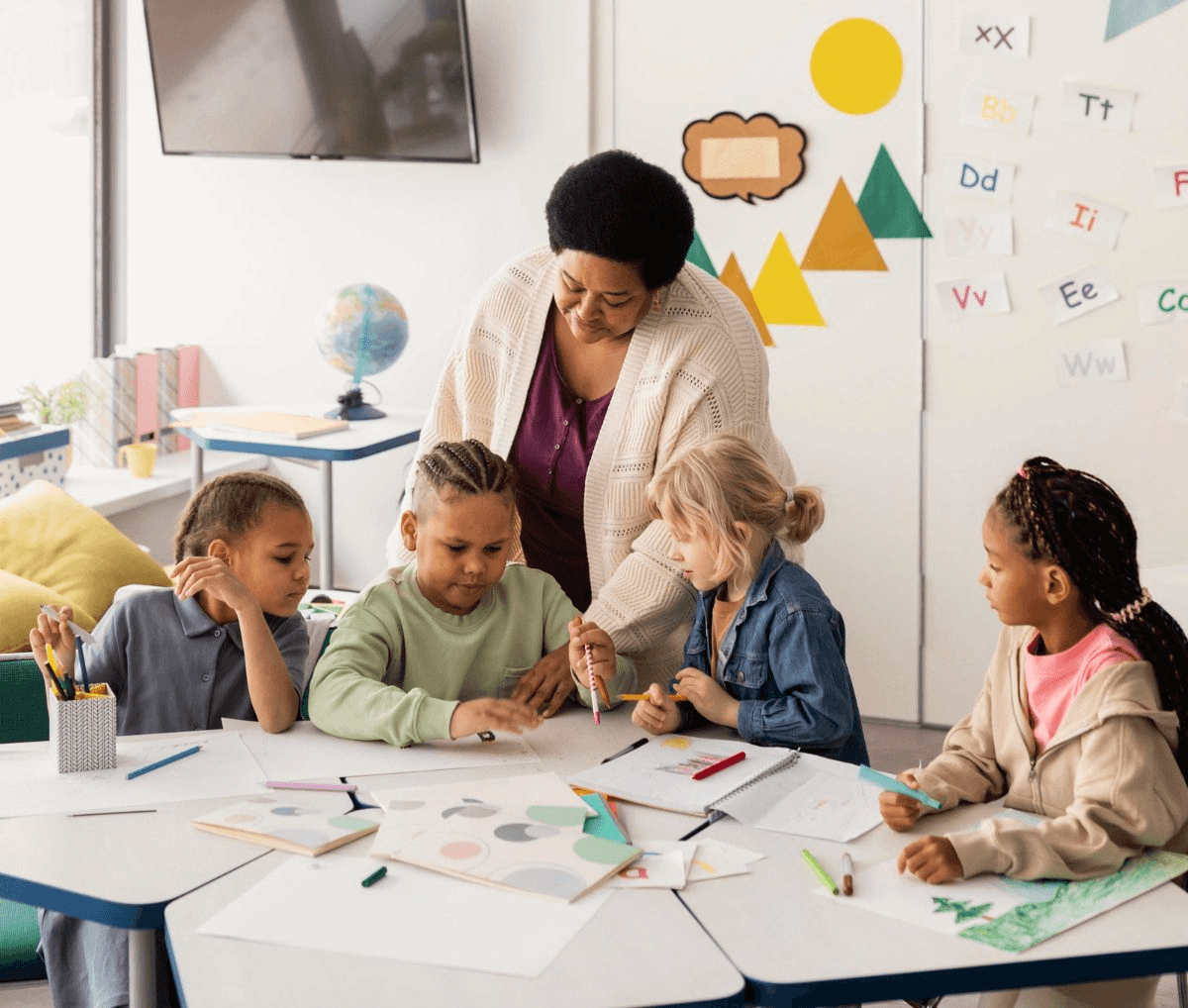 A teacher leaning over a classroom table, guiding four young students as they draw and color
